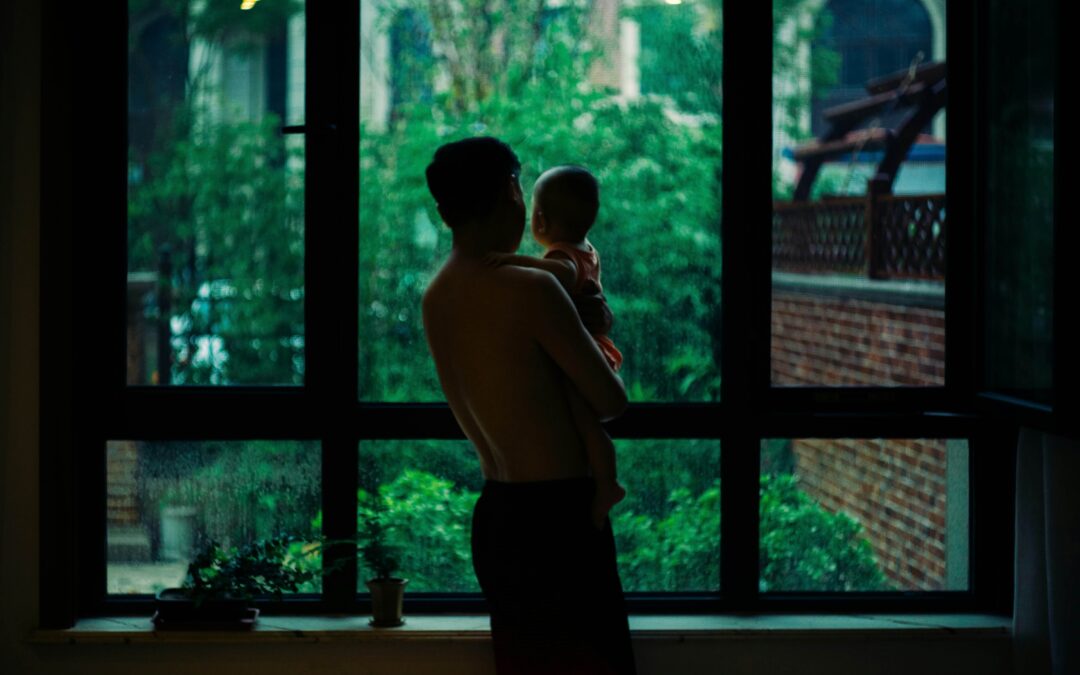 topless man carrying baby standing beside window.